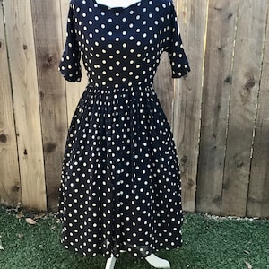 May include: A navy blue dress with white polka dots. The dress has a scoop neckline, short sleeves, and a fitted bodice that flows into a flared skirt. The dress is displayed on a white mannequin against a wooden fence background.