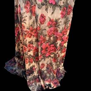 May include: A sheer fabric with a vibrant floral pattern. The design features large, red and pink roses with green leaves against a beige background. The fabric drapes, showcasing the floral print and a dark, watercolor-like border.