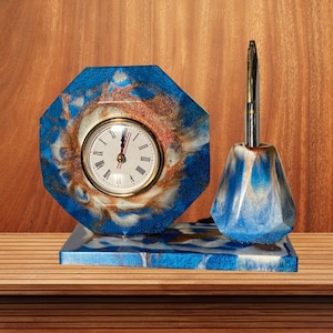May include: A blue and white resin clock and pen holder set. The clock face is white with black Roman numeral hour markers and black hands. The pen holder is a blue and white resin geometric shape.