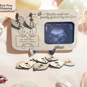 May include: Wooden countdown calendar with an elephant design and a frame for an ultrasound image. The calendar has hearts with numbers 1-20 and the text "Weeks until our family grows by two feet". The frame has the text "Baby" and the text "Our Baby Girl is Expected to Arrive 02.02.2025".