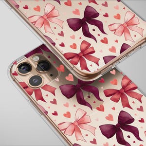 Puede incluir: Dos fundas transparentes para teléfono con un patrón repetido de lazos y corazones de acuarela en tonos rosa y burdeos. Las fundas están sobre un fondo beige claro.