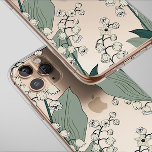 Può includere: Due custodie trasparenti per telefono con un design floreale. Le custodie presentano un motivo di fiori bianchi di mughetto e foglie verdi su sfondo beige. Le custodie sono angolate, mostrando il design e la fotocamera del telefono.