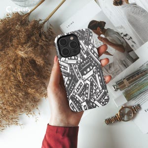 May include: A white iPhone case with a black and white pattern of cassette tapes. The case has a camera cutout and a raised lip around the edges.