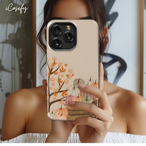 May include: A beige phone case with a fall-themed design. The case features a tree with orange leaves and a pile of pumpkins in various shades of orange, white, and green. The case has a cutout for the phone's camera lens.