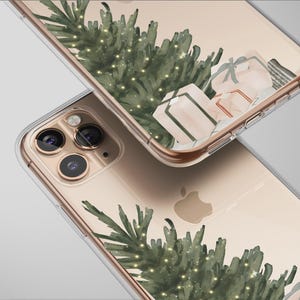 May include: Clear phone case featuring a watercolor design of a green Christmas tree with tiny lights and wrapped gifts. The case is shown on a gold-colored phone, with the Apple logo visible. The design has a festive holiday theme.
