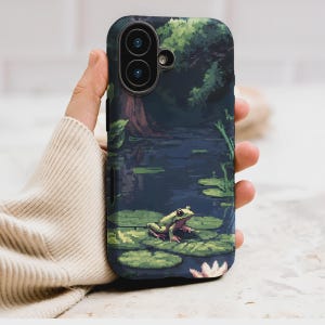 May include: A pixelated phone case featuring a serene pond scene. The design includes a green frog on lily pads, a dark blue pond, and a backdrop of trees. The case has a dark blue border and a camera cutout.
