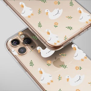May include: Two clear phone cases with a repeating pattern of white cartoon ducks, yellow flowers, and green leaves on a beige background. The cases are angled, showcasing the design and the phone's camera.
