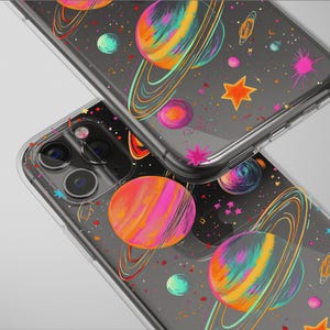 May include: Clear phone cases with a colorful space-themed design. The cases feature planets with rings, stars, and splashes of pink, orange, and blue on a dark background. The cases are shown stacked.