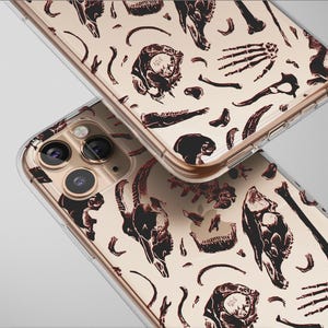 May include: Two clear phone cases with a repeating pattern of black skulls and bones on a beige background. The cases are angled, showing the camera lenses and the edges of the phones.