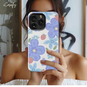 May include: A white phone case with a floral pattern featuring blue flowers and red strawberries. The case has a clear back and a black camera cutout.