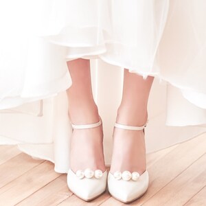 May include: White pointed-toe heels with ankle straps and pearl embellishments. The shoes are worn under a white dress, creating a bridal or formal look. The heels have a stiletto design.