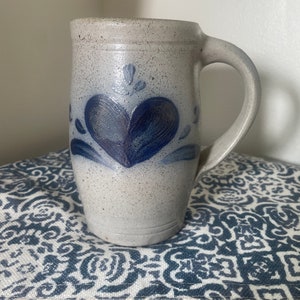 May include: A gray stoneware mug with a blue heart design. The mug has a handle and is decorated with a blue heart and swirls.