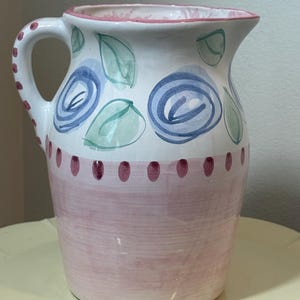 May include: A ceramic pitcher featuring a white upper section decorated with hand-painted blue roses and green leaves. The lower section is painted in a solid light pink with a row of dark pink dots along the rim where the colors meet.
