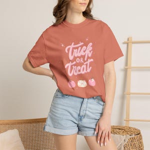 May include: A terracotta-coloured short-sleeve t-shirt featuring the text "Trick or Treat" in a light pink script font. Below the text are three small, illustrated pumpkins in shades of pink and white, accented with small star shapes.