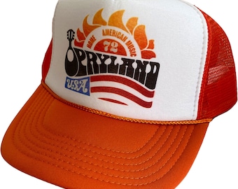 May include: A white and orange trucker hat with a red mesh back. The front of the hat features a logo with the text "Home of American Music '72" and "OPRYLAND USA".