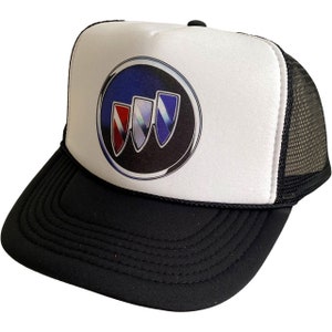 Puede incluir: Una gorra de camionero blanca y negra con una parte trasera de malla negra. La parte delantera de la gorra presenta un gran logotipo de Buick colorido.