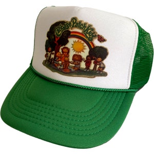 May include: A green and white trucker hat with a Cabbage Patch Kids logo and a rainbow. The logo features a group of Cabbage Patch Kids dolls playing in a park.