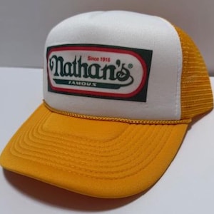 以下が含まれることがあります： 黄色と白のトラッカーハット。前面にNathan's Famousのロゴがあり、つばとメッシュバックは黄色です。ロゴは赤、緑、白のアクセントが付いた長方形の中にあります。「Since 1916」の文字がロゴの上にあります。