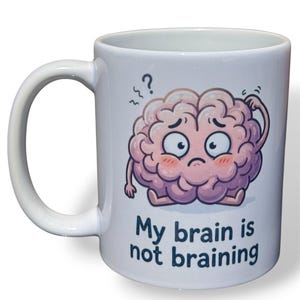May include: A white ceramic mug featuring a cartoon illustration of a pink brain character with a confused expression, scratching its head. Above the brain is a question mark, and below it is the text "My brain is not braining" in dark blue.