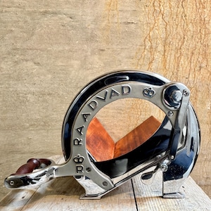 Puede incluir: Un cortador de pan vintage plateado con una tabla de cortar de madera. El cortador tiene un mango y una palanca. El nombre de la marca "R.B. RADVAD" está impreso en el lateral.