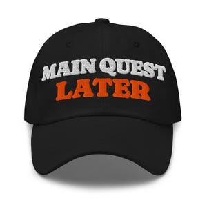 May include: A black baseball cap featuring embroidered text on the front. The words MAIN QUEST are stitched in white thread, while the word LATER is stitched in orange thread below it. The cap has a curved brim and a structured crown design.