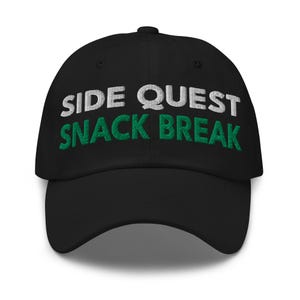 Könnte beinhalten: Eine schwarze Baseballkappe mit gesticktem Text auf der Vorderseite. Die obere Zeile zeigt SIDE QUEST in weißem Garn, die untere Zeile SNACK BREAK in grünem Garn. Die Kappe hat einen gebogenen Schirm und eine strukturierte Krone mit Ösen.