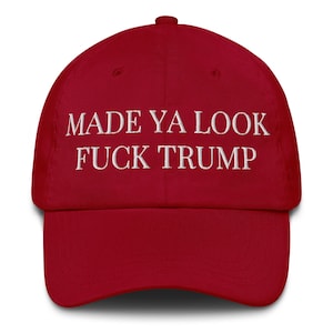 May include: A red baseball cap with the text "MADE YA LOOK FUCK TRUMP" embroidered in white. The cap has a curved peak and a button on top. This hat is a political statement.