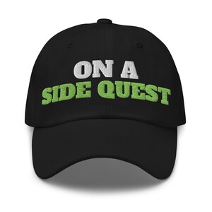 May include: A black baseball cap featuring embroidered text on the front. The words ON A are stitched in white, while the words SIDE QUEST are stitched in a bright lime green color. The cap has a curved brim and a structured six-panel crown design.