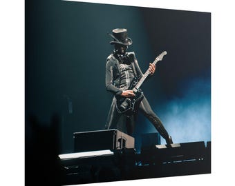 Pode incluir: Um artista em um traje preto com cartola e máscara toca uma guitarra elétrica preta no palco. O palco está escuro com iluminação azul e fumaça, criando uma atmosfera dramática. A roupa do artista inclui um casaco longo e calças justas.