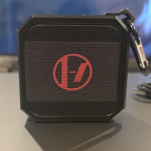 May include: A black, square-shaped portable speaker with a red logo on the front. The speaker has a mesh front and a black carabiner clip attached to the top. The logo is a stylized design within a circle.