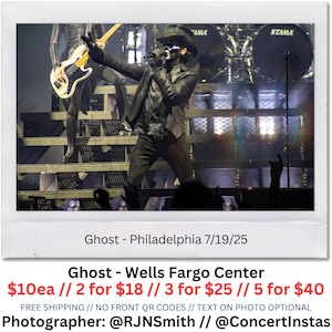 May include: A live concert photo of the band Ghost performing on stage. The lead singer, wearing a black outfit and a mask, is gesturing towards the audience. The stage features drums and a bass guitar. Text on the photo reads "Ghost - Philadelphia 7/19/25".