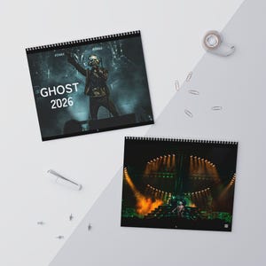 May include: Two spiral-bound calendars featuring concert photography. One calendar displays the text "GHOST 2026" over an image of a performer on stage. The second calendar shows a stage with bright lights. The calendars are on a white surface.