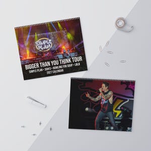 May include: A 2027 wall calendar featuring the Simple Plan Bigger Than You Think Tour. The cover displays a concert stage with the band name and tour details. The second page shows a musician performing on stage with a microphone and tattoos.