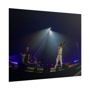 Puede incluir: Escena de concierto en vivo con dos artistas en el escenario. Uno viste un atuendo blanco y guantes rojos, con el brazo levantado. El otro lleva una camiseta negra y toca el teclado. La iluminación del escenario crea una atmósfera dramática.