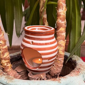 May include: A white and brown striped ceramic oil burner with a round opening for a tea light. The burner is sitting on a brown surface with green plants in the background.