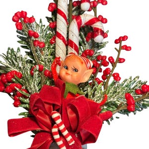 Op de afbeelding: Een kerstdecoratie met een kleine elfenfiguur met een gestreepte muts, twee rood-witte zuurstokken, trossen helderrode bessen en groene dennentakken bestrooid met witte sneeuw, afgewerkt met een grote rode fluwelen strik.