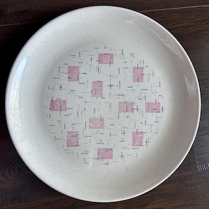 Może przedstawiać: Okrągły, kremowy ceramiczny talerz z nakrapianą powierzchnią. Talerz posiada centralny wzór różowych kwadratów i czarnych linii, tworząc geometryczny wzór. Talerz nadaje się do serwowania potraw.