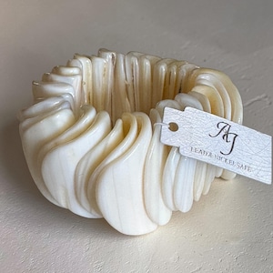 Puede incluir: Una pulsera gruesa de color crema con un diseño ondulado y en capas. La pulsera tiene un acabado brillante y está hecha de un material tallado. Una etiqueta adjunta a la pulsera dice "Lead & Nickel Safe".