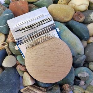 May include: A silver knitting loom with a wooden base, surrounded by smooth, colorful stones.