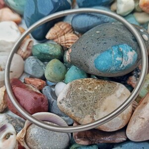 May include: A silver metal spring ring bracelet lying on a bed of colorful stones and seashells.
