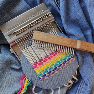 May include: A close-up of a loom with a rainbow-colored woven tapestry. The loom is made of metal and has a wooden comb. The tapestry is made of yarn and is woven in a simple pattern.