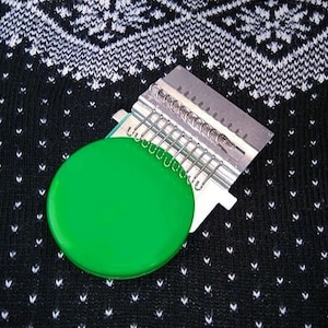 May include: A green circular knitting loom with a silver metal knitting needle attachment. The loom is on a black and white patterned fabric.