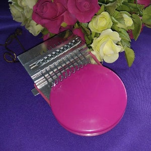 May include: A pink kalimba, a thumb piano, with metal tines and a round, pink resonator. The instrument is set against a purple background with a bouquet of roses in shades of pink, red, and yellow.