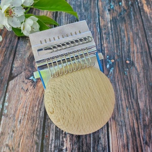 May include: A silver knitting loom with a wooden base. The loom has a series of metal hooks and a spring-loaded mechanism.