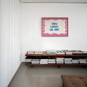 May include: A wooden shelf unit with two shelves filled with books. A pink framed artwork with the text "HOW LUCKY WE ARE" hangs on the white wall behind the shelf.
