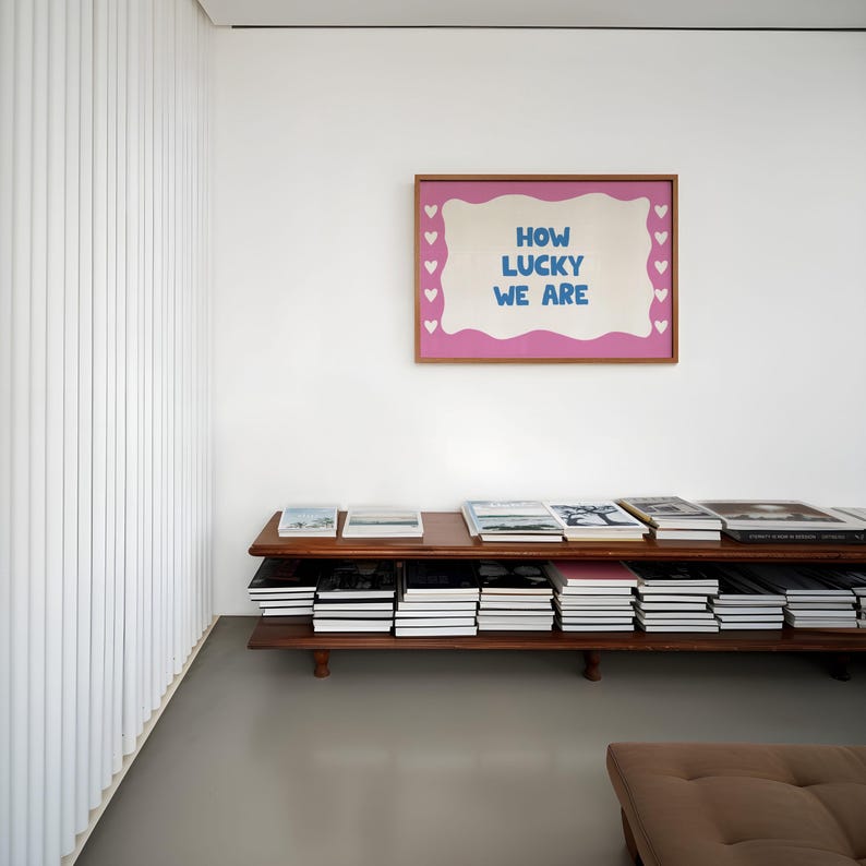May include: A wooden shelf unit with two shelves filled with books. A pink framed artwork with the text "HOW LUCKY WE ARE" hangs on the white wall behind the shelf.