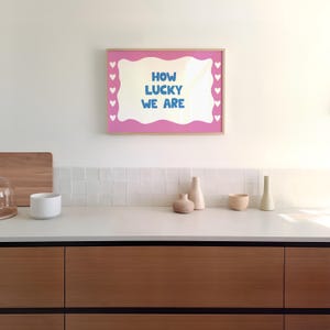 May include: A pink framed print with a white wavy border and white hearts around the edge. The print says "How lucky we are" in blue text.