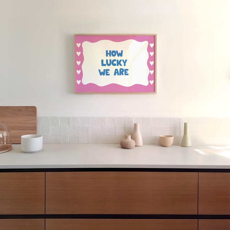 May include: A pink framed print with a white wavy border and white hearts around the edge. The print says "How lucky we are" in blue text.