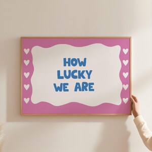 May include: A pink framed print with a white background and a blue text overlay that reads "HOW LUCKY WE ARE". The print has a white heart border.