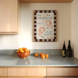 May include: A framed print with a brown and blue checkered border. The print features a white background with the text "An Americano and a Flat White Please" in black and orange lettering. Two cartoon hands hold coffee cups.